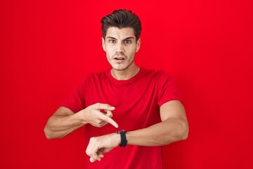 Young hispanic man standing over red background in hurry pointing to watch time, impatience, upset...