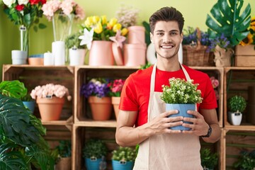 Young hispanic man working at florist shop holding plant looking positive and happy standing and smiling with a confident smile showing teeth