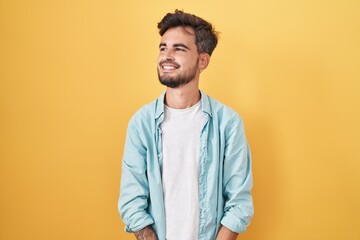Young hispanic man with tattoos standing over yellow background looking away to side with smile on face, natural expression. laughing confident.
