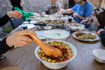 The wife fills a salad bowl with a large bowl of yogurt next to it to provide the guests with what they need at lunchtime at the large collective table for the large family.