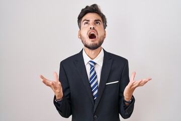 Young hispanic man with tattoos wearing business suit and tie crazy and mad shouting and yelling with aggressive expression and arms raised. frustration concept.
