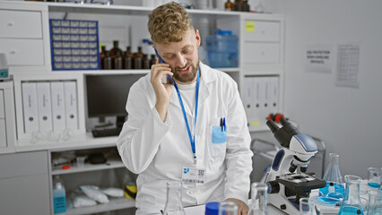 A young, caucasian, bearded man in a laboratory, wearing a lab coat and talking on the phone...