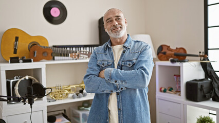 Smiling middle-aged bald man with beard in a denim jacket standing in a music studio with...