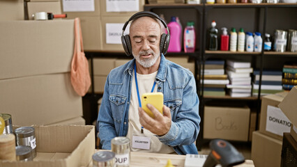 Mature man with headphones using smartphone in a warehouse full of donations and supplies.