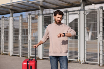 Fototapeta na wymiar Being late. Worried man with red suitcase looking at his watch outdoors