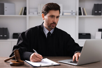 Fototapeta premium Judge with gavel and laptop writing in papers at wooden table indoors