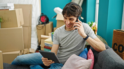 Young hispanic man talking on smartphone using touchpad sitting on sofa at new home