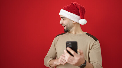 Young arab man wearing christmas hat using smartphone over isolated red background