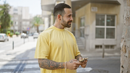 A bearded hispanic man in casual wear stands outdoors in the city, holding a smartphone while looking to the side.