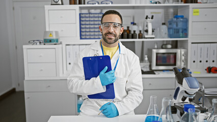Naklejka na ściany i meble A young hispanic man with a beard poses in a laboratory setting, wearing safety glasses and a lab coat while holding a clipboard.