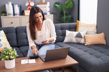 Young beautiful hispanic woman using laptop counting dollars at home