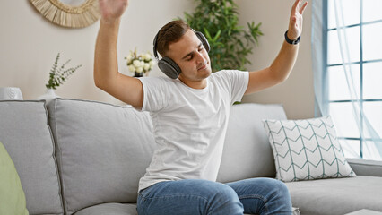 A joyful young hispanic man relaxes with music on headphones, dancing on a sofa in a cozy living room setting.