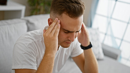 A young adult man looking stressed indoors, possibly experiencing a headache or distress, with a neutral bedroom backdrop.