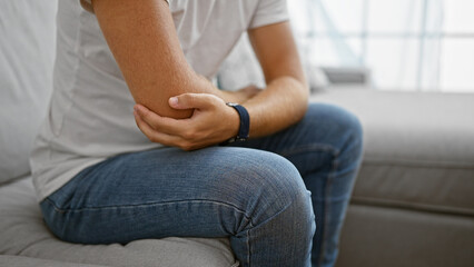 Fototapeta na wymiar A young hispanic man experiencing elbow pain while sitting on a couch indoors.