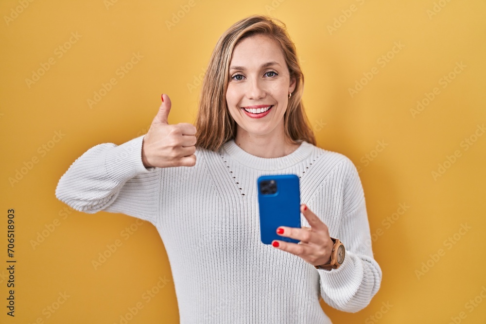 Sticker Young blonde woman using smartphone typing message doing happy thumbs up gesture with hand. approving expression looking at the camera showing success.