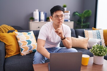Young asian man using laptop at home sitting on the sofa thinking concentrated about doubt with...