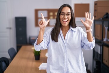 Young hispanic woman at the office showing and pointing up with fingers number seven while smiling...
