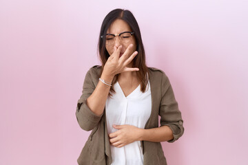 Middle age chinese woman wearing glasses over pink background smelling something stinky and disgusting, intolerable smell, holding breath with fingers on nose. bad smell