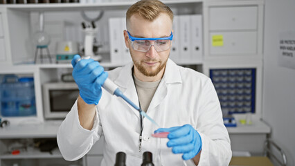 A focused young man in labcoat performs an experiment in a laboratory, pipetting a blue substance...