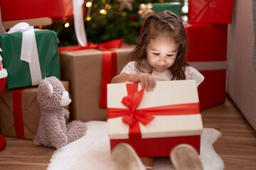 Fototapeta na wymiar Adorable hispanic girl unpacking gift sitting on floor by christmas tree at home