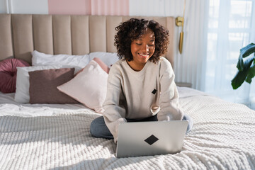 African American girl using laptop in bed at home office typing chatting reading writing email....