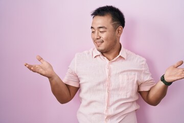 Fototapeta premium Chinese young man standing over pink background smiling showing both hands open palms, presenting and advertising comparison and balance