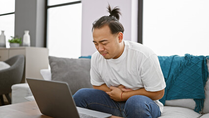Handsome young chinese man using laptop on sofa, visibly suffering from sharp stomachache at home