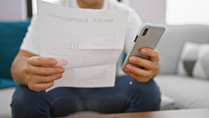 In his living room, expression etched on his face, a man, sitting on the sofa, has his hands busy holding and using his cellphone to type a message about his electrical bill.