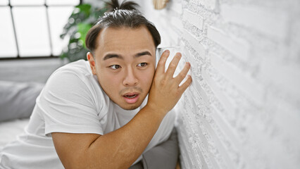 Nosy young chinese man sneakily listening through bedroom wall with glass, overcome by curiosity...