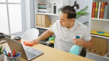 Dedicated young chinese man confidently springs into action at work, cleaning office table with sprayer and cloth