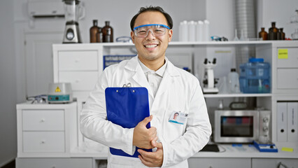 Smiling young chinese scientist, a charming professional man working with confidence and security in the lab, clipboard in hand - obrazy, fototapety, plakaty