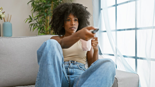 African american woman watching tv sitting on sofa drinking coffee at home