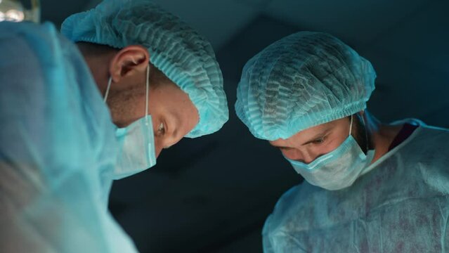 Close-up pair of Caucasian doctors in surgical uniforms working in operating room. Focused surgeons operating patient standing on both sides of him in modern hospital emergency department, slow motion