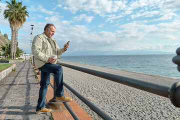 Smiling middle-aged bearded man drinking coffee and using smartphone on seaside in sunny winter or spring day.