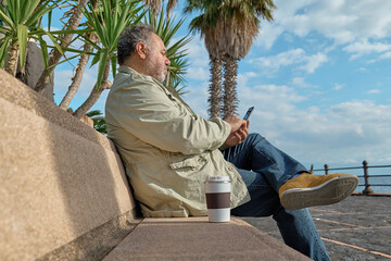 Smiling middle-aged bearded man using smartphone while sitting on the bench on seaside in sunny...