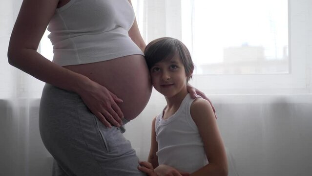 A pregnant woman in a white T-shirt stands at the window with a curtain, her eldest son of six years old. Boy strokes, kisses, hugs the belly of a pregnant mother.