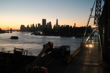 Traffic driving over the Williamsburg Bridge at night with the New York financial district skyline and other bridges in the background at sunset.