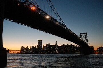 Williamsburg Bridge at night with the New York financial district skyline in the background at sunset.  Statue of Liberty is visible in the far distance.