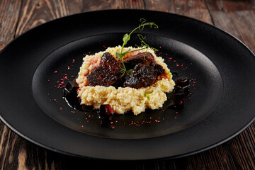 Braised beef cheeks with millet on a black plate, served on a rustic wooden table