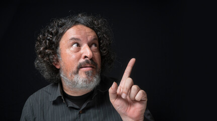 Man with white beard and black curly hair with funny expression, pointing up with his hand, wearing black shirt against black background