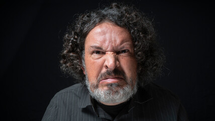 Man with white beard and black curly hair with angry expression, looking straight at camera, wearing black shirt against black background