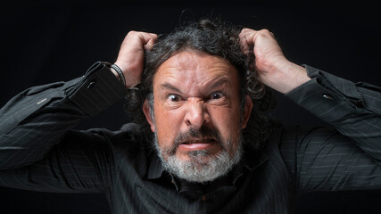 Man with white beard and black curly hair with very angry expression, grabbing his hair with hands, wearing black shirt against black background