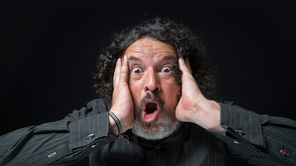 Man with white beard and black curly hair with surprised expression, hands on face, wearing black shirt against black background