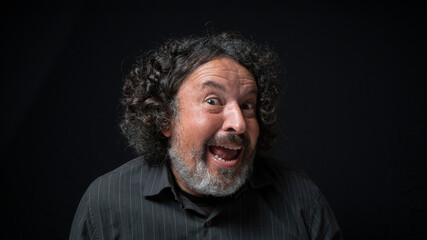 Man with white beard and black curly hair with happy and funny expression, eyes looking at camera, wearing black shirt against black background