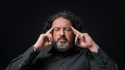 Man with white beard and black curly hair with concentrated expression, with his fingers on his temples in his hand, wearing black shirt against black background