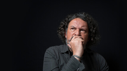 Man with white beard and black curly hair with doubtful and angry expression, with his hand on his face, wearing black shirt against black background