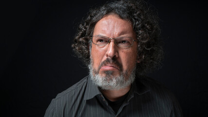 Man with white beard and black curly hair with frustrated expression, grimacing with mouth, wearing black shirt against black background