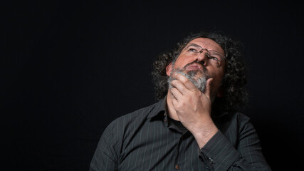 Man with white beard and black curly hair with expression of indecision and doubt, looking up, wearing black shirt against black background