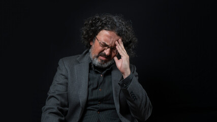 Man with white beard and black curly hair with pained expression, with his hand on his forehead, wearing black shirt against black background