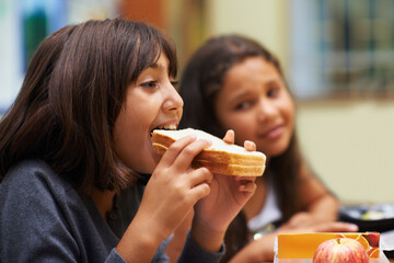 Hungry girl, student and eating sandwich in classroom at school for meal, break or snack time. Young kid, person or elementary child biting bread for lunch, fiber or nutrition in class during recess - Powered by Adobe